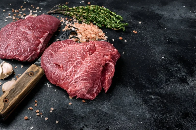 Raw beef cheek meat on a wooden cutting board with herbs.